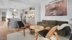 Uma sala de estar moderna com um sofá verde, cadeiras de madeira e uma mesa de centro redonda. Uma cozinha é visível ao fundo, com armários brancos e uma ilha central.