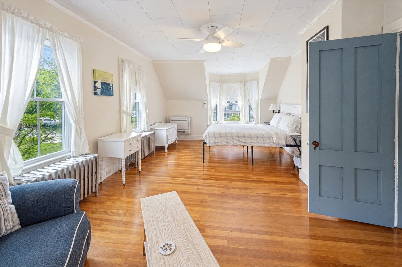 Ein Schlafzimmer mit Bett, Schreibtisch, Heizkörper, einem fenster mit Vorhängen, einem Deckenventilator und einer Tür.