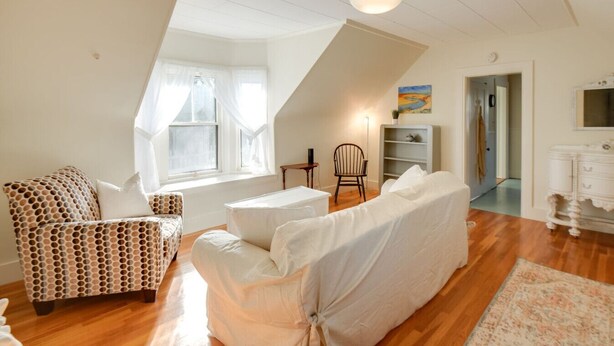 A spacious loft bedroom with a high ceiling, a large window with sheer curtains, a cozy seating area with patterned furniture, and a classic dresser.