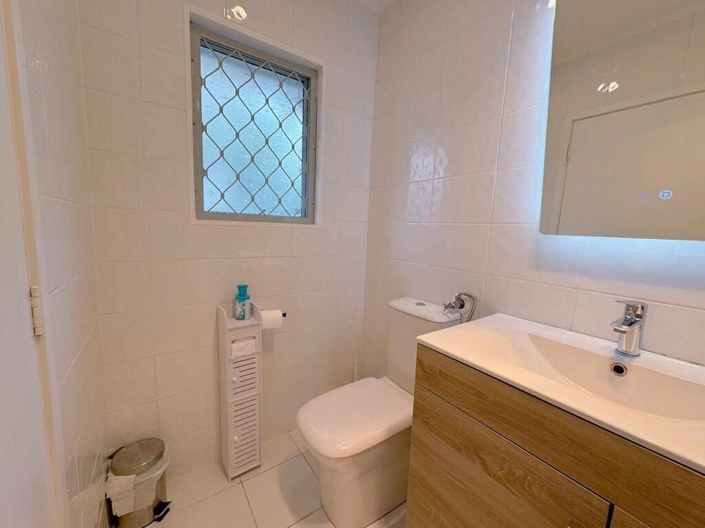 Een badkamer met witte tegels, een toilet, een wastafel met spiegel en een raam met metalen tralies.