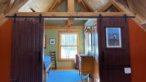 Interior rústico de cabana com teto alto, vigas de madeira e estrutura central de madeira. Há uma porta que dá para um quarto com cama e janela.