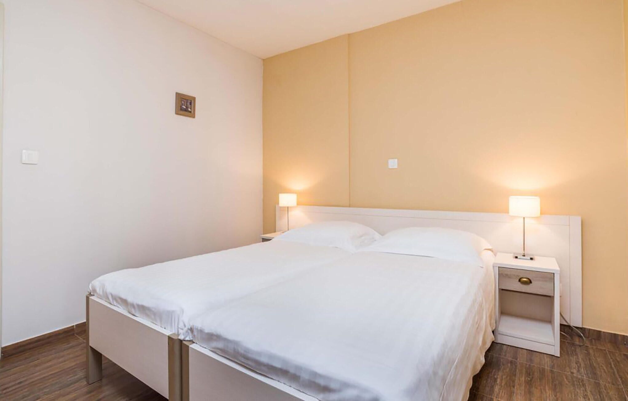 A neatly made bed with white linens in a hotel room. There are two bedside tables with lamps, one on each side of the bed. The room has a simple, clean design with light-colored walls and wooden flooring.