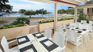 Un área de comedor al aire libre con sillas y mesas blancas, vistas a un lago y plantas en macetas.