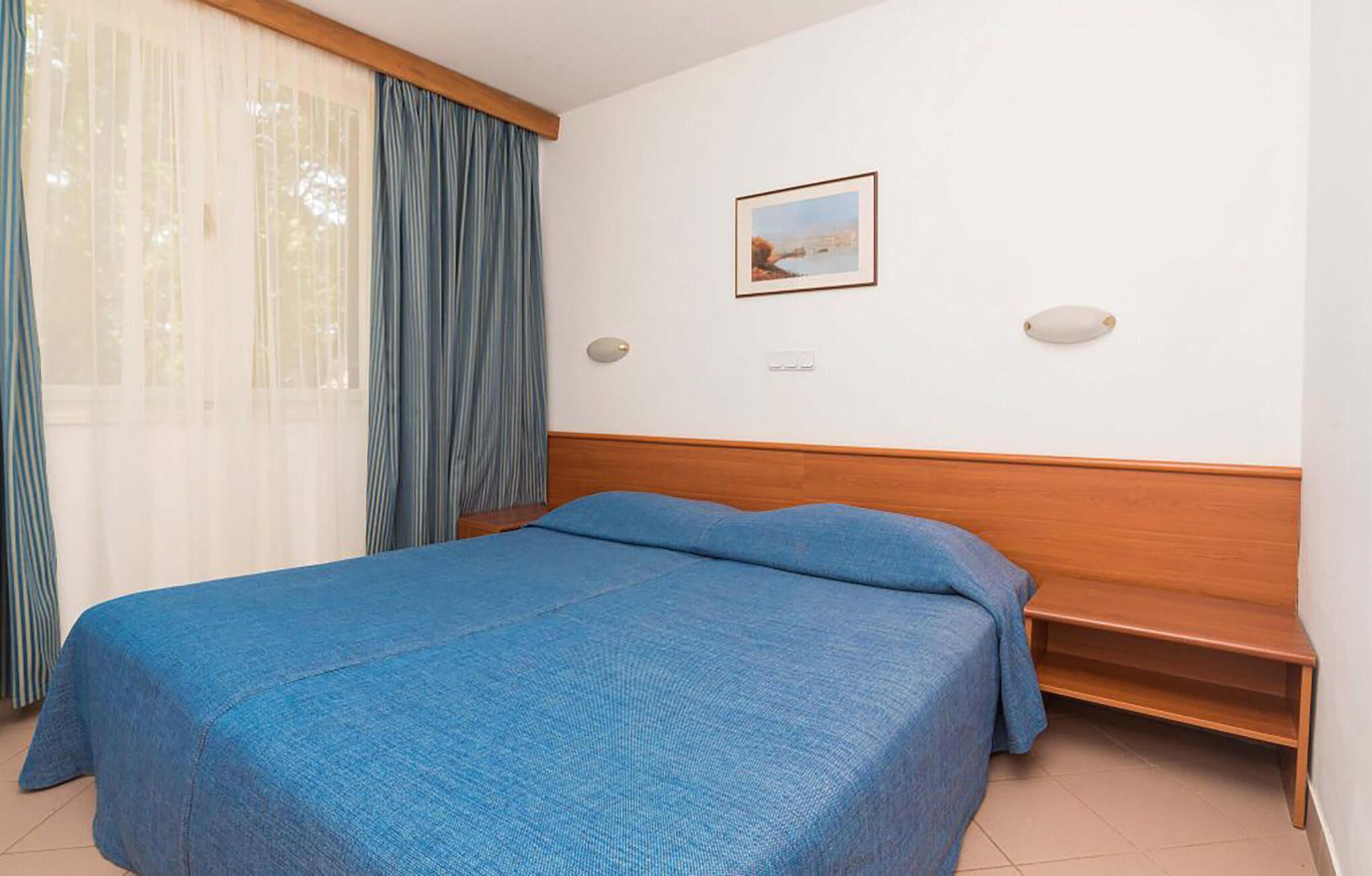 A bedroom with a bed, a wooden headboard, a bench, a window with curtains, and a framed picture on the wall.