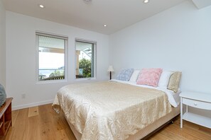 A bedroom with a large bed, two windows, a bedside table, and a lamp. - Premier Oceanfront Luxury in Oak Bay South (Oak Bay South)