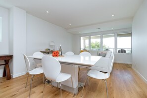 A modern dining area with a white table and chairs, a wooden floor, and a view through large windows. - Premier Oceanfront Luxury in Oak Bay South (Oak Bay South)