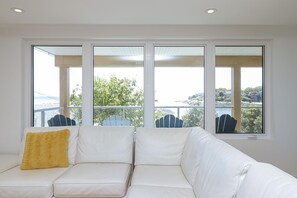 A white leather sofa with a yellow pillow, large windows with a view of a balcony and trees, and a white ceiling. - Premier Oceanfront Luxury in Oak Bay South (Oak Bay South)