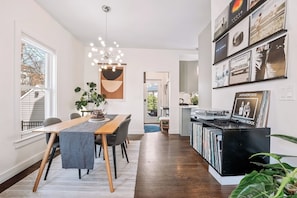 A modern kitchen with a built-in CD rack, a dining area with a wooden table and chairs, and a large window with blinds. - Luxury Boston Base · 2 Suites · 2 Baths · Red Line · Parking · Davis Square (Somerville)