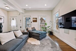 A modern living room with a sectional sofa, a flat-screen TV, and large windows. - Luxury Boston Base · 2 Suites · 2 Baths · Red Line · Parking · Davis Square (Somerville)