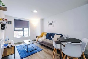 Ein modernes Wohnzimmer mit einer Couch, einem Couchtisch, einem Esstisch mit Stühlen, einem großen Fenster und einer Stehlampe.