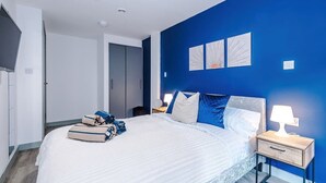 Quarto moderno com uma cama grande, mesa de cabeceira com abajur e obra de arte na parede azul.