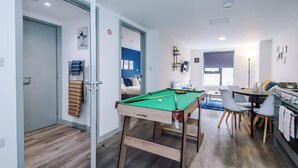 Quarto moderno com mesa de bilhar, área de refeições com cadeiras, cozinha com fogão e cama com cabeceira azul.