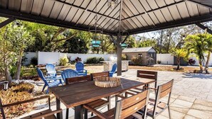 Une terrasse extérieure avec une table et des chaises en bois, un espace salon couvert et un petit jardin agrémenté de plantes et d’une clôture blanche.