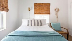 Une chambre à coucher comprenant un lit, une chaise, une lampe fixée au mur, une fenêtre avec des stores et une décoration murale en macramé.