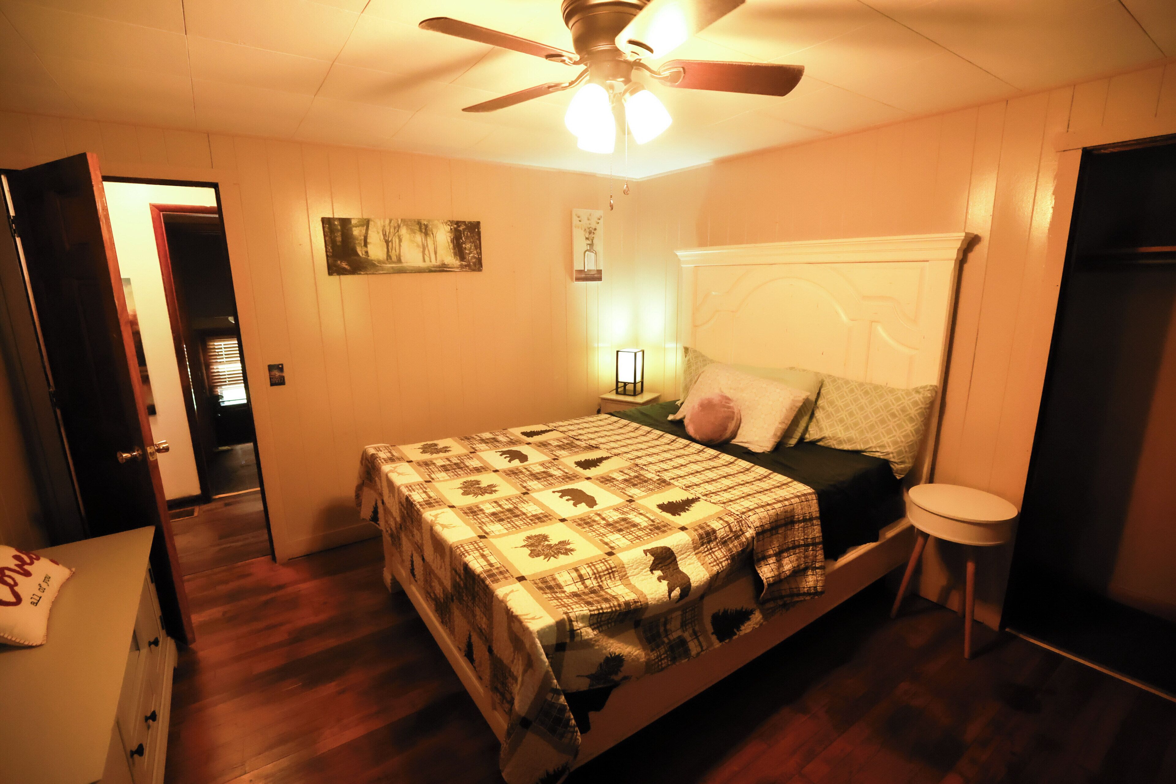 A bedroom with a bed, a ceiling fan, a nightstand, a bedside table, and a door leading to another room.