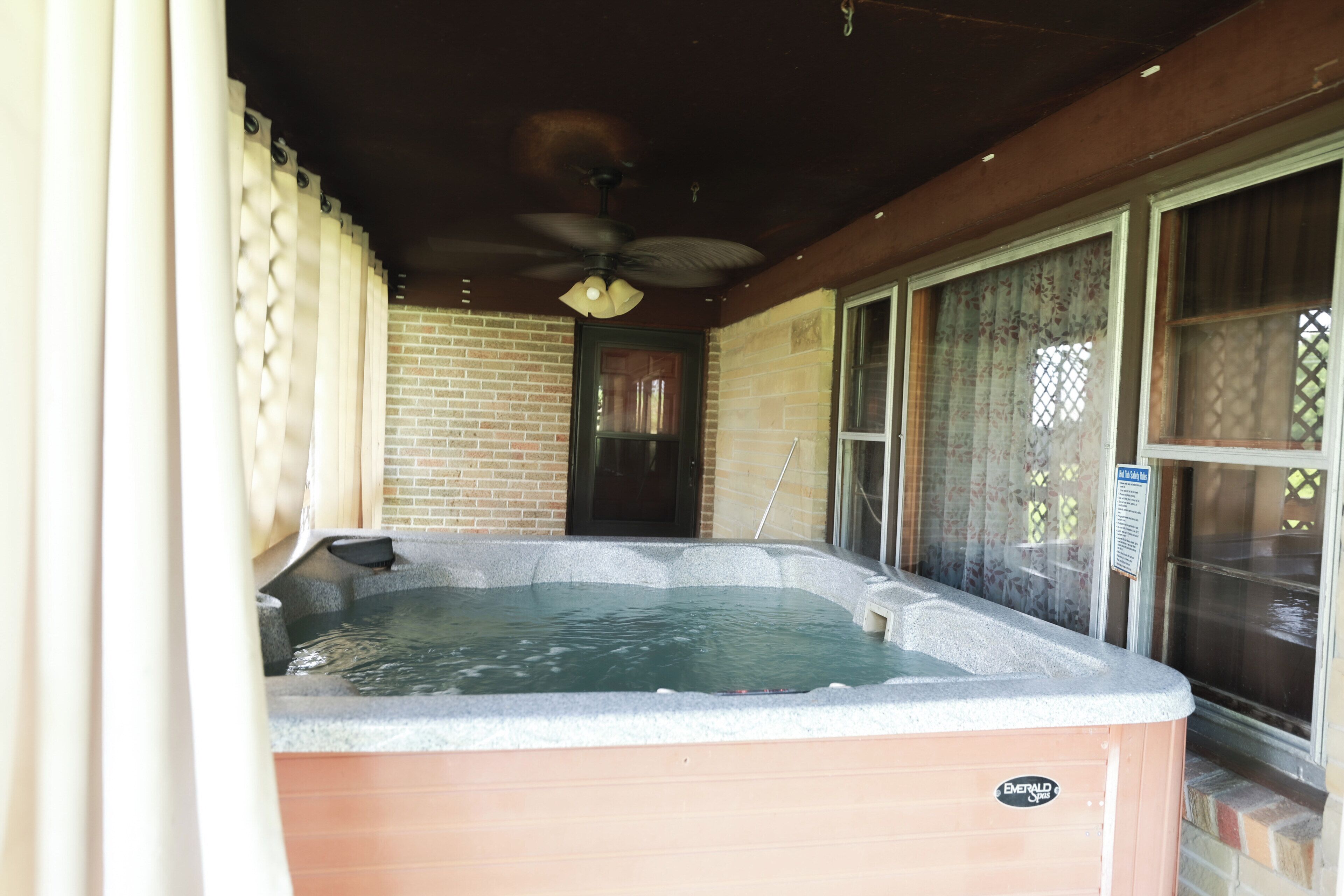 Un bain à remous avec un ventilateur de plafond, un mur en briques et une fenêtre ornée d’un rideau à motifs floraux.