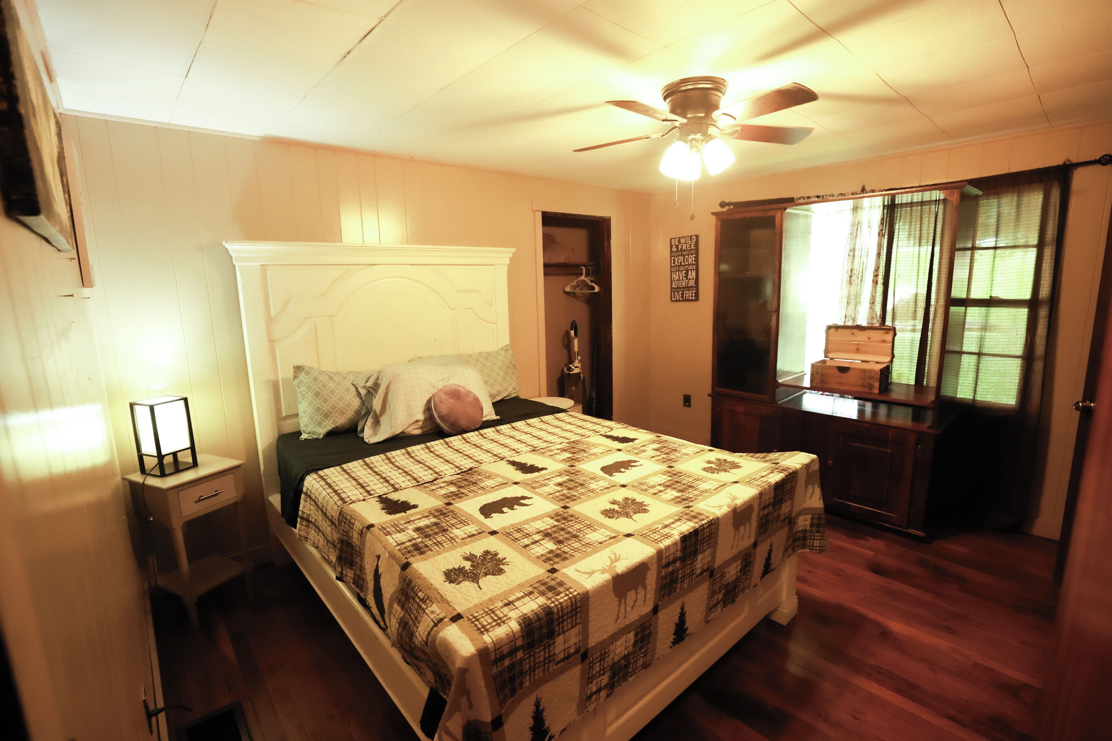 Une chambre à coucher comprenant un lit, une table de chevet, un ventilateur de plafond, une commode et une fenêtre avec des rideaux.