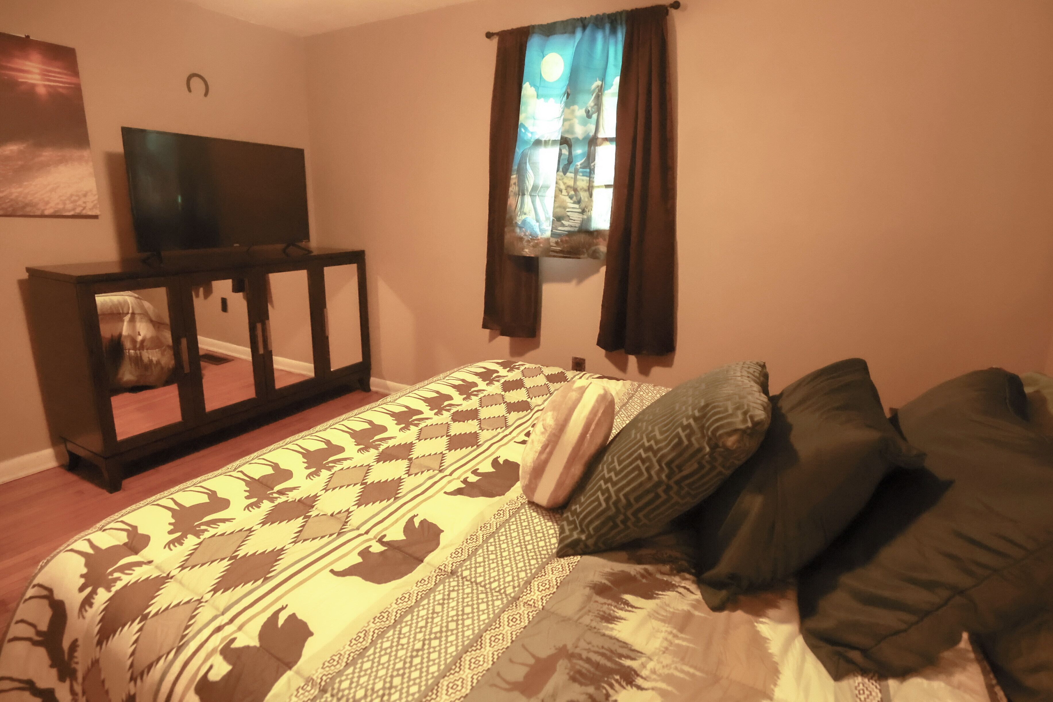 Un dormitorio con una cama, un televisor sobre un soporte, una ventana con cortinas y una obra de arte colgada en la pared.