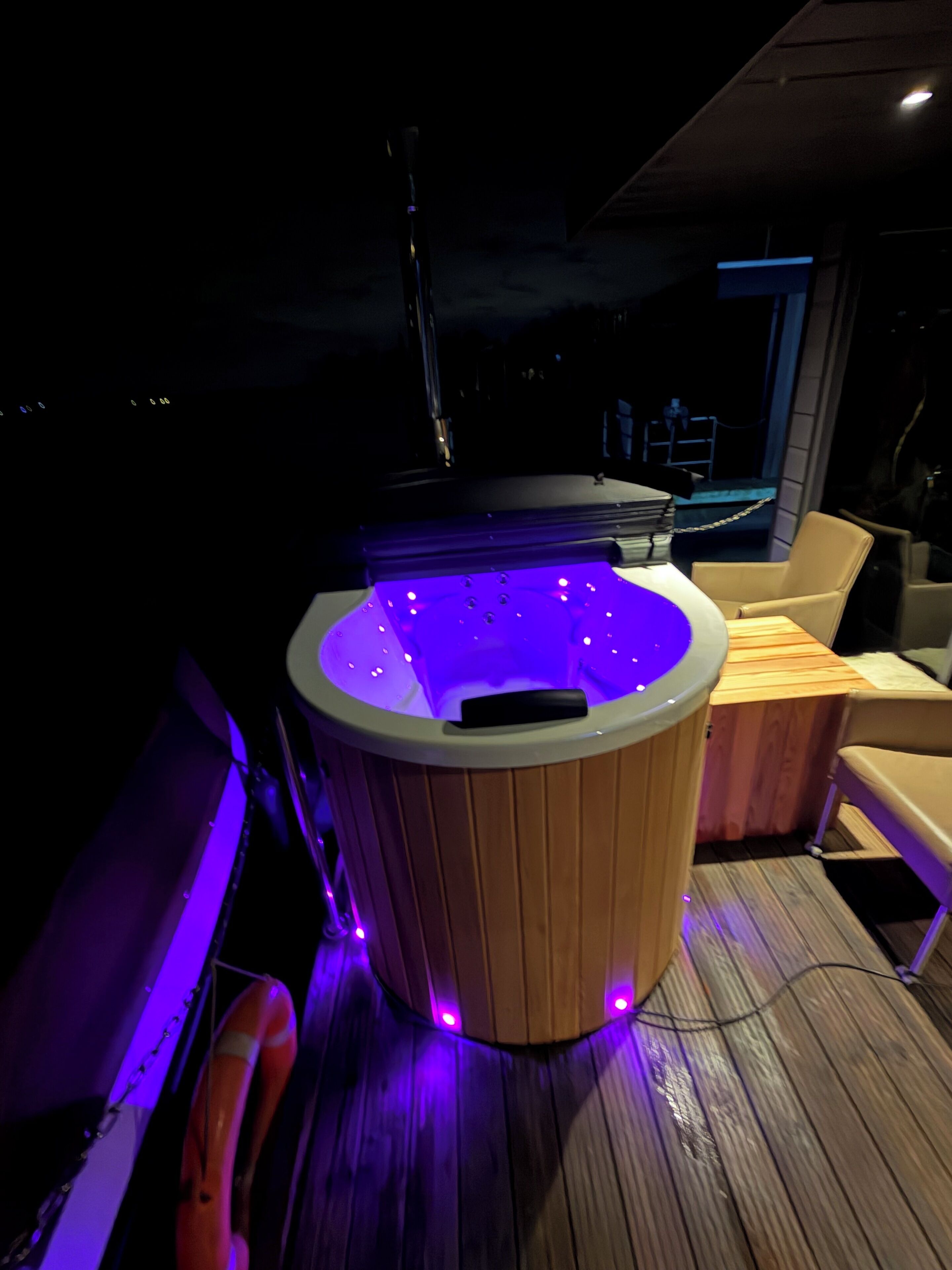 A hot tub with a wooden exterior and purple lighting, located on a wooden deck with a chair and a table nearby.