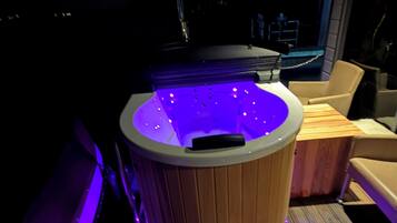 A hot tub with a wooden exterior and purple lighting, located on a wooden deck with a chair and a table nearby.