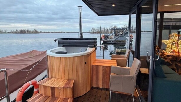 A modern outdoor deck with a hot tub, wooden seating, and a glass enclosure overlooking a body of water. - Exclusive Houseboat 12 x 5 m with sauna and hot tub (Maasbommel)