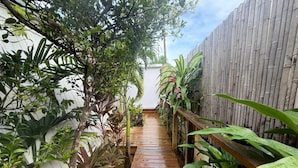 Un sendero de madera bordeado por exuberante vegetación y una valla de bambú, que conduce a un edificio blanco.
