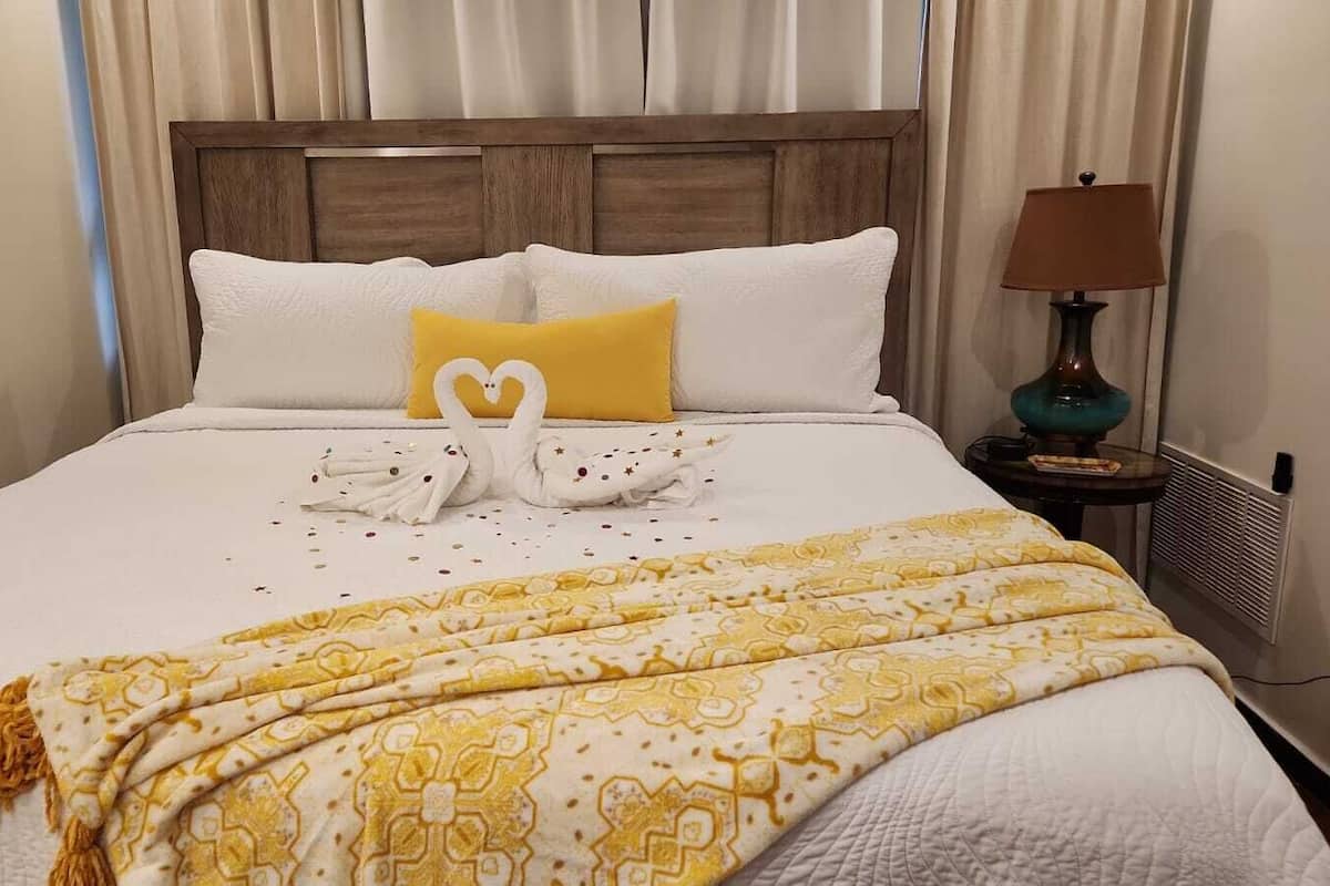 A neatly made bed with a white and yellow patterned bedspread, a heart-shaped towel arrangement, and a yellow pillow.