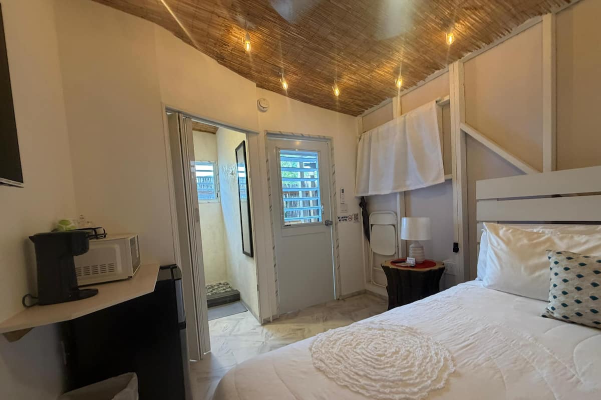 A bedroom with a bed, a bedside table with a lamp, a microwave, and a small trash bin.