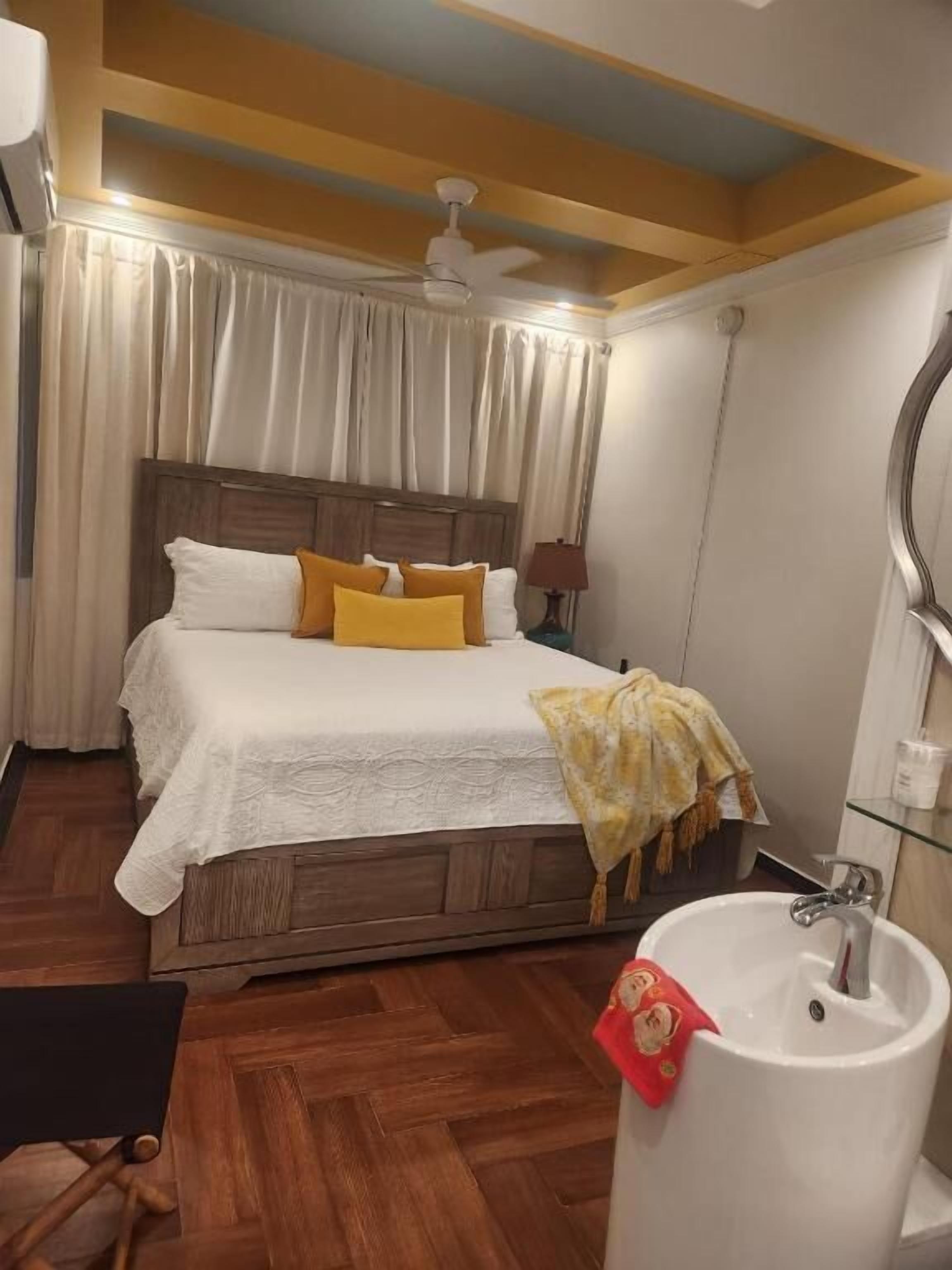 Habitación de hotel con una cama de madera, ropa de cama blanca, ventilador de techo, lámpara de noche y baño con lavamanos y espejo.