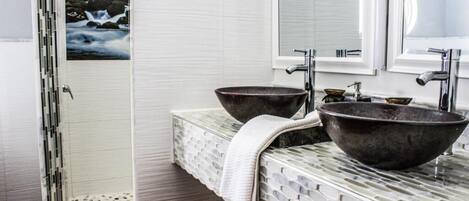 Un baño con dos lavamanos grandes y oscuros, una zona de ducha con azulejos blancos y una imagen de cascada en la pared.