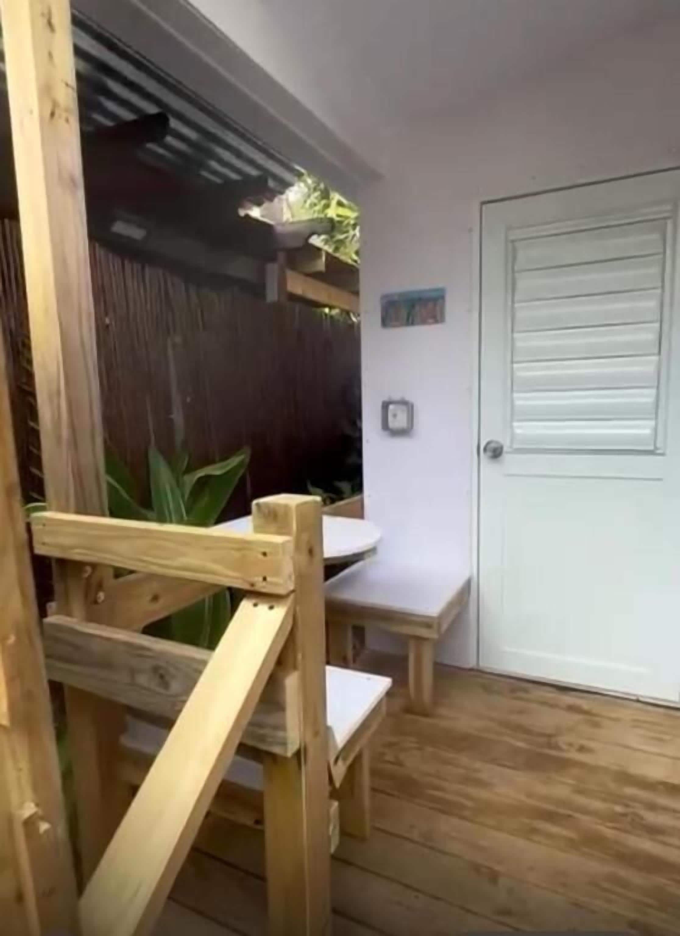 Um deck de madeira com um banco e uma pequena mesa, uma porta branca e uma cerca de madeira.
