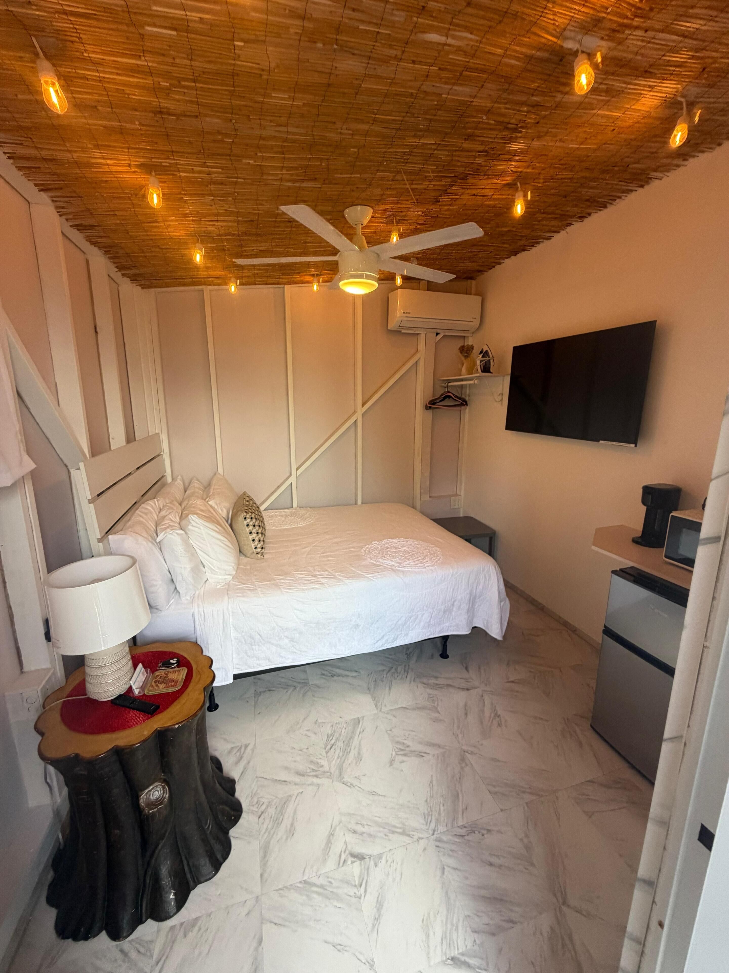 A bedroom with a bed, bedside table, lamp, ceiling fan, air conditioning unit, TV, and a small kitchenette.