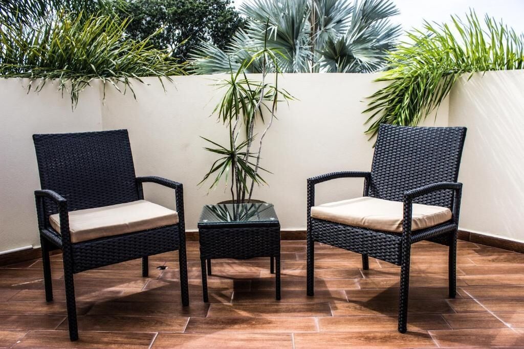 Deux fauteuils en osier avec des coussins, une petite table en verre et une plante en pot, le tout installé dans un espace patio extérieur doté d’un sol carrelé et d’un mur blanc.
