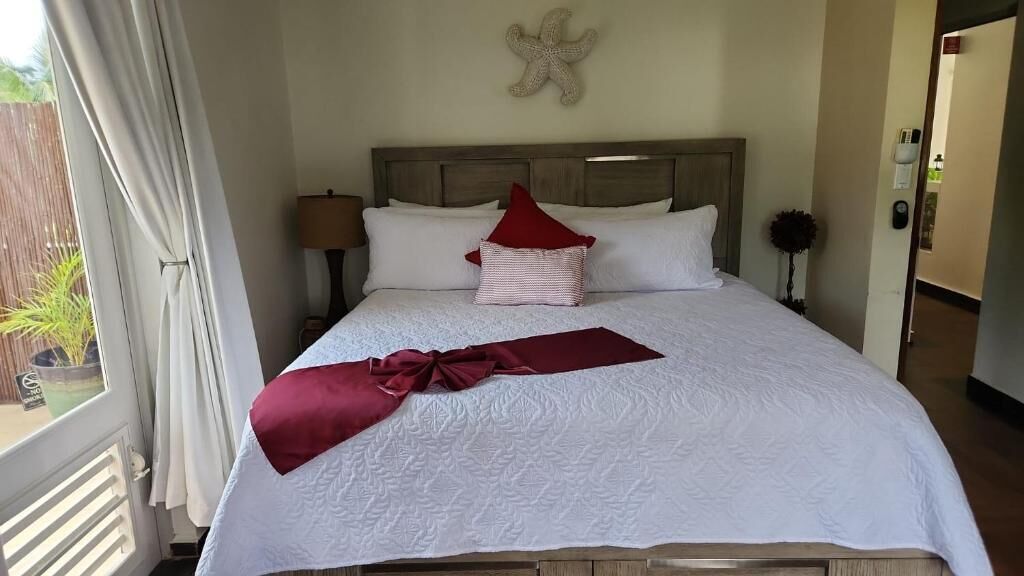 A neatly made bed with white bedding and two red pillows, a starfish decoration on the wall, and a view of the outdoors through a window.