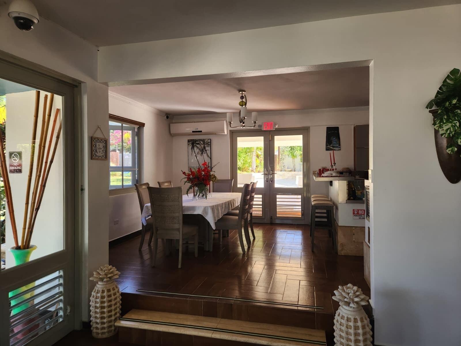 A dining area with a table set for four, surrounded by chairs. There is a door leading outside, a wall-mounted air conditioning unit, and a security camera above the door.
