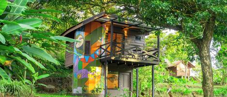 A treehouse with a colorful mural, surrounded by lush greenery and a clear sky.