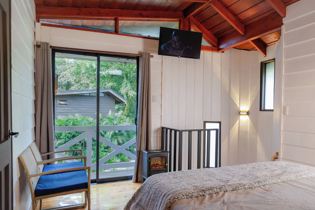 Een slaapkamer met een bed, een televisie, een open haard, een balkon met uitzicht op groen, en een houten plafond.