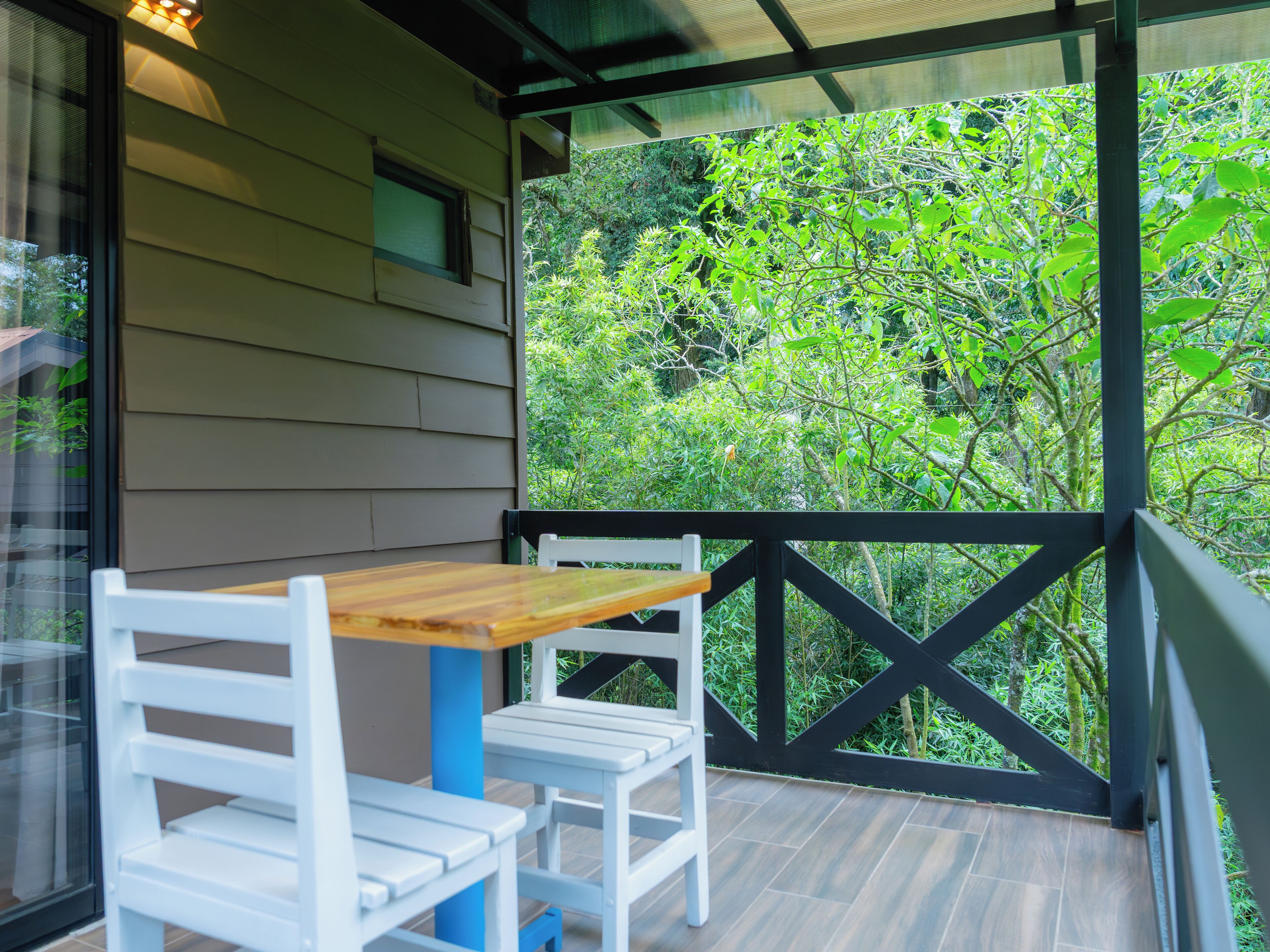 A covered outdoor seating area with a wooden table and white chairs, overlooking a lush garden.