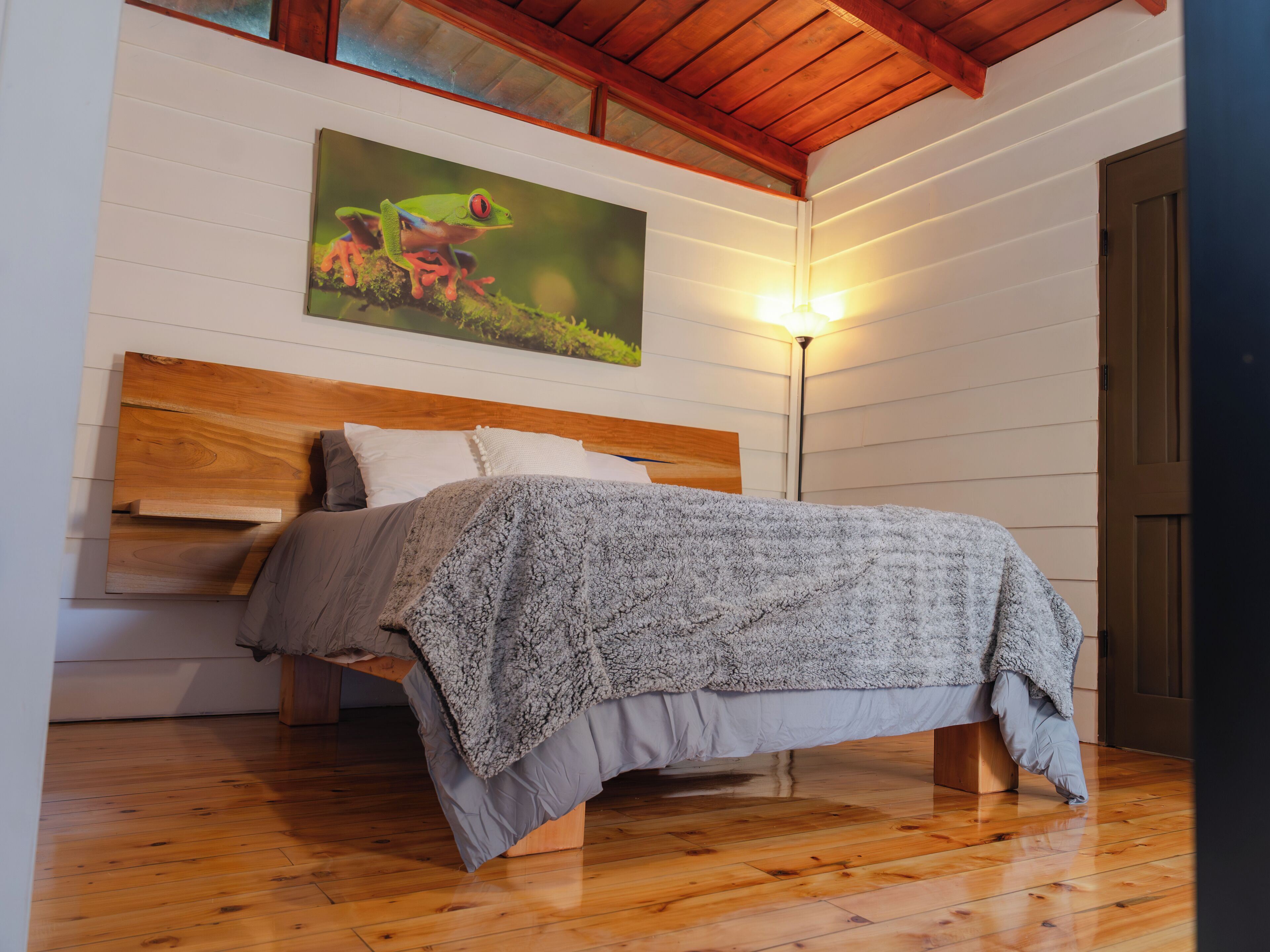 A bedroom with a wooden headboard, a bed with a grey blanket, a floor lamp, and a painting of a frog on the wall.