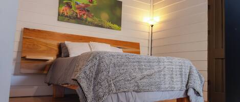 A bedroom with a wooden headboard, a bed with a grey blanket, a floor lamp, and a painting of a frog on the wall.