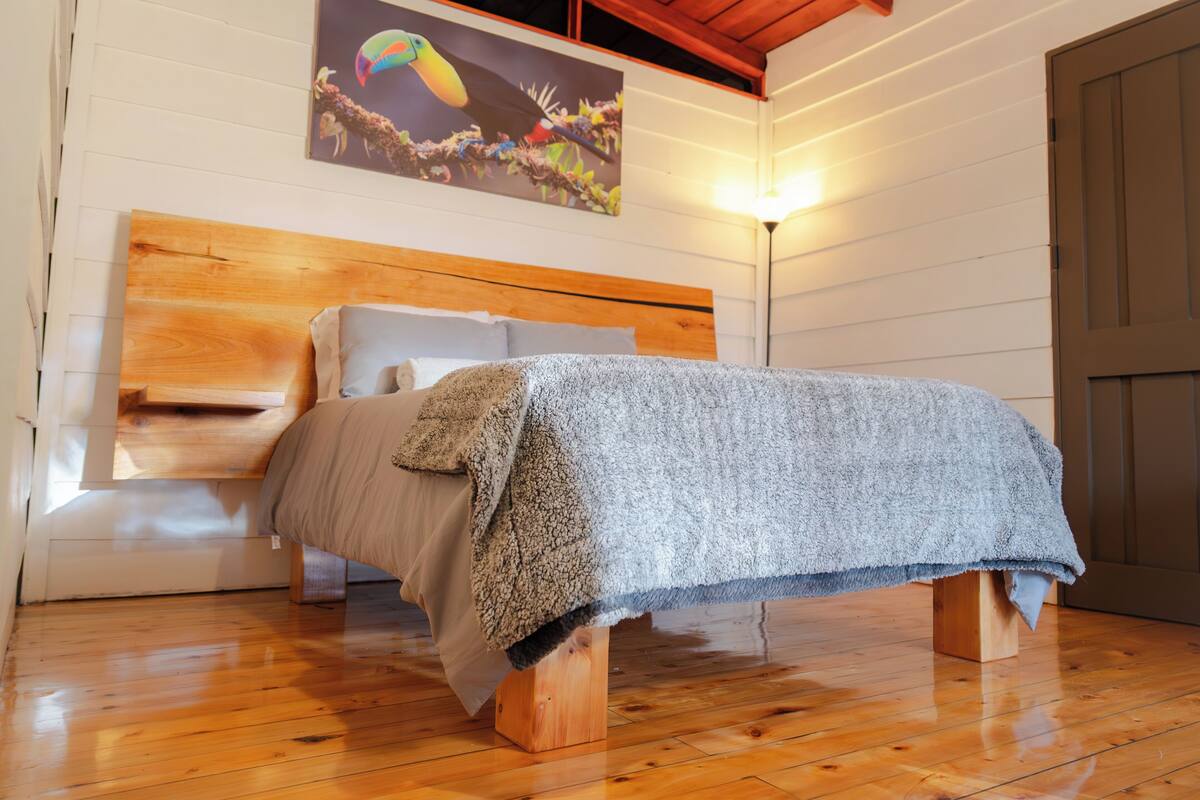 Een bed met houten frame, hoofdeinde, een grijze deken en een nachtkastje van hout.
