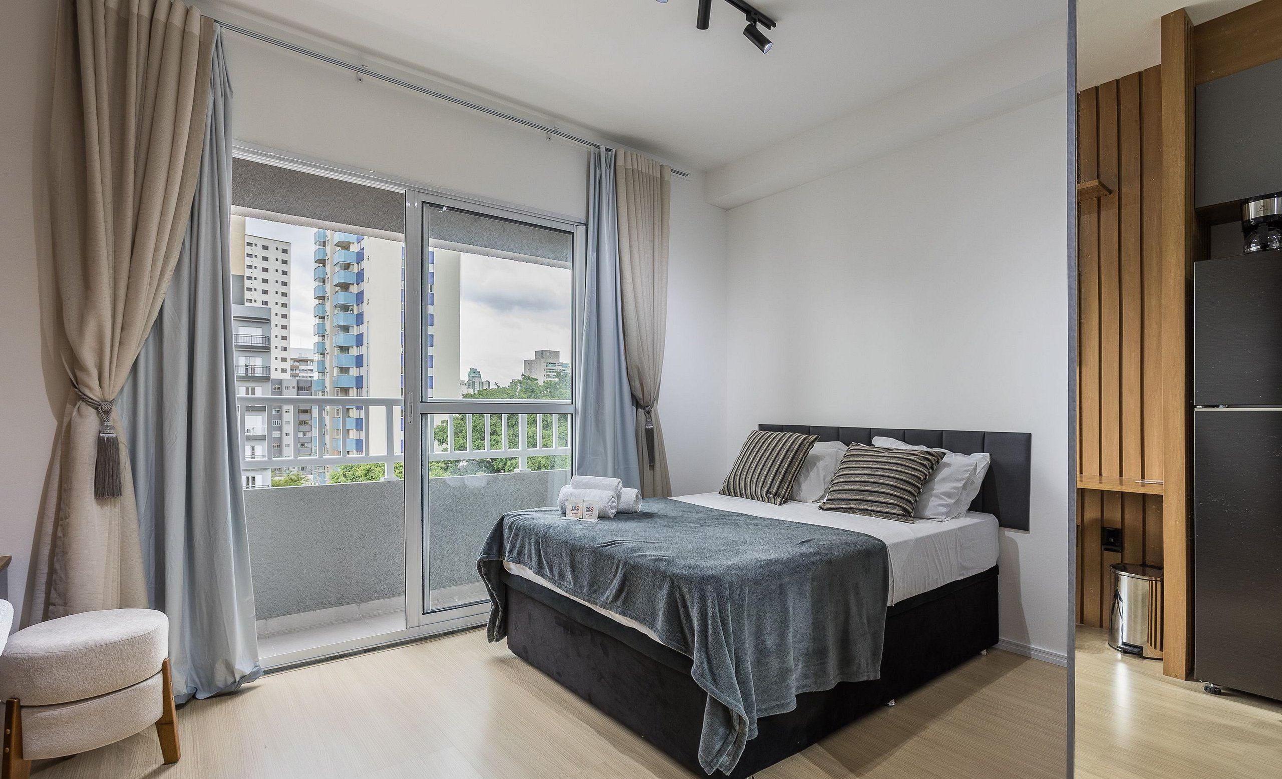 A modern bedroom with a bed, curtains, a view of buildings, and a wooden cabinet.