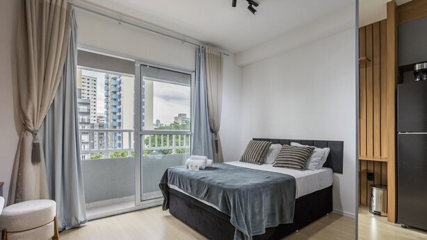 Une chambre moderne avec un lit, des rideaux, une vue sur des immeubles et une armoire en bois.