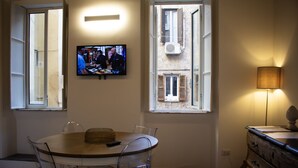 Ein Essbereich mit rundem Tisch, Stühlen, einem an der Wand montierten Fernseher und einer Lampe auf einer Kommode.
