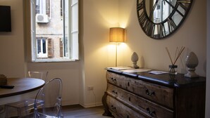 Ein Zimmer mit einem Holzkleiderschrank, einer runden Wanduhr, einer Lampe, einem Fenster mit Fensterläden und einem klaren Stuhl.