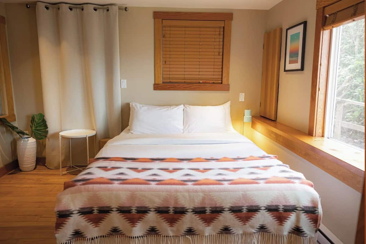Um quarto com cama, janela com persianas, mesa de cabeceira, abajur e uma planta.