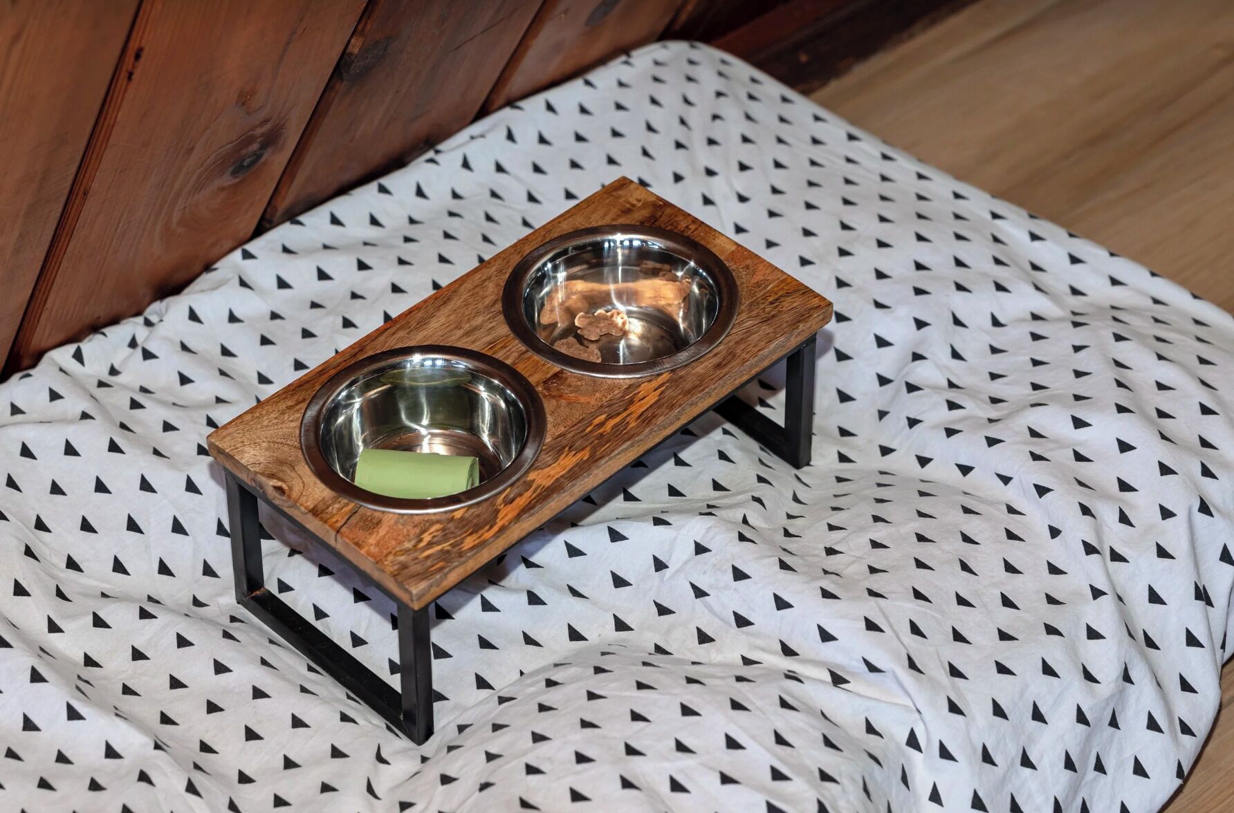 A wooden pet feeding station with two bowls on a patterned fabric.