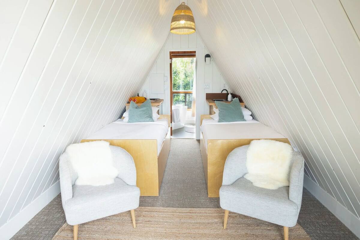 Quarto sótão aconchegante em formato triangular, com duas camas, uma janela grande e um lustre.
