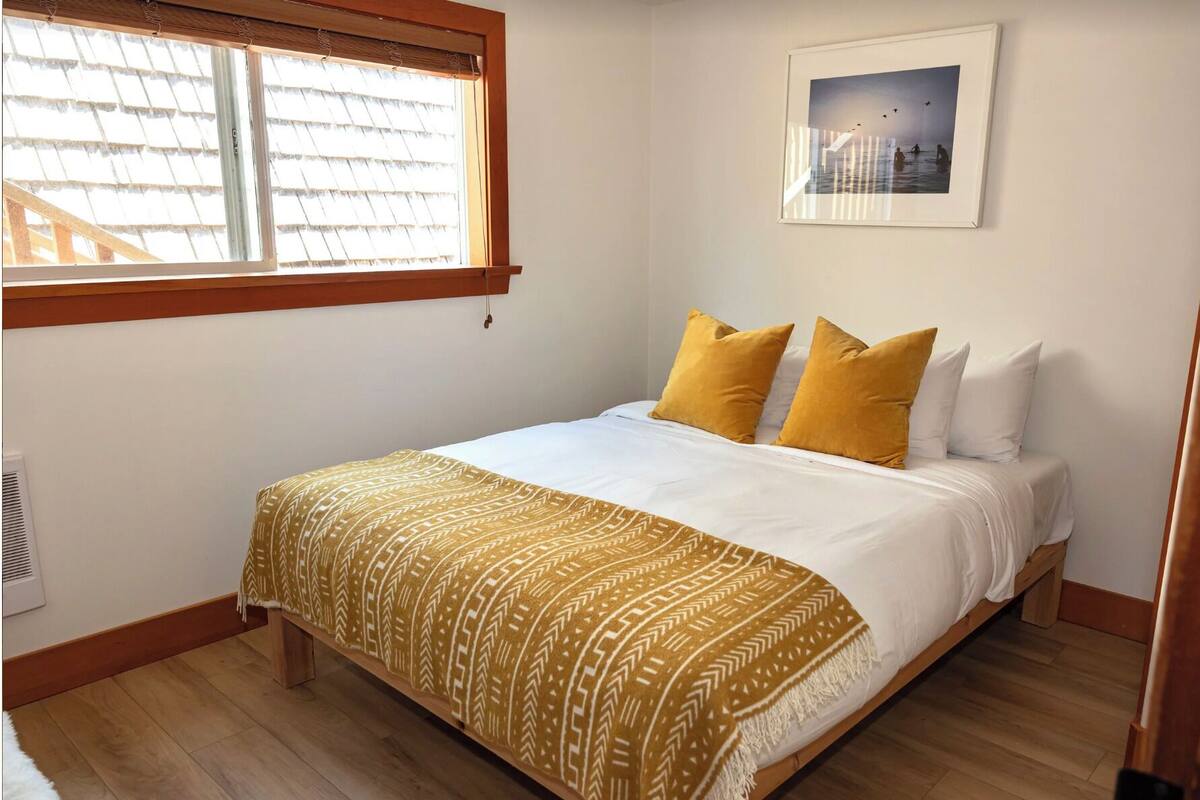 Quarto com cama, janela com persianas, quadro na parede e manta decorativa.