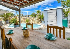 A wooden dining table with green plates and bowls, a woven basket, and a stone wall with plants in the background. - Paradera Breeze - 2-BR Vacation Home (Paradera)